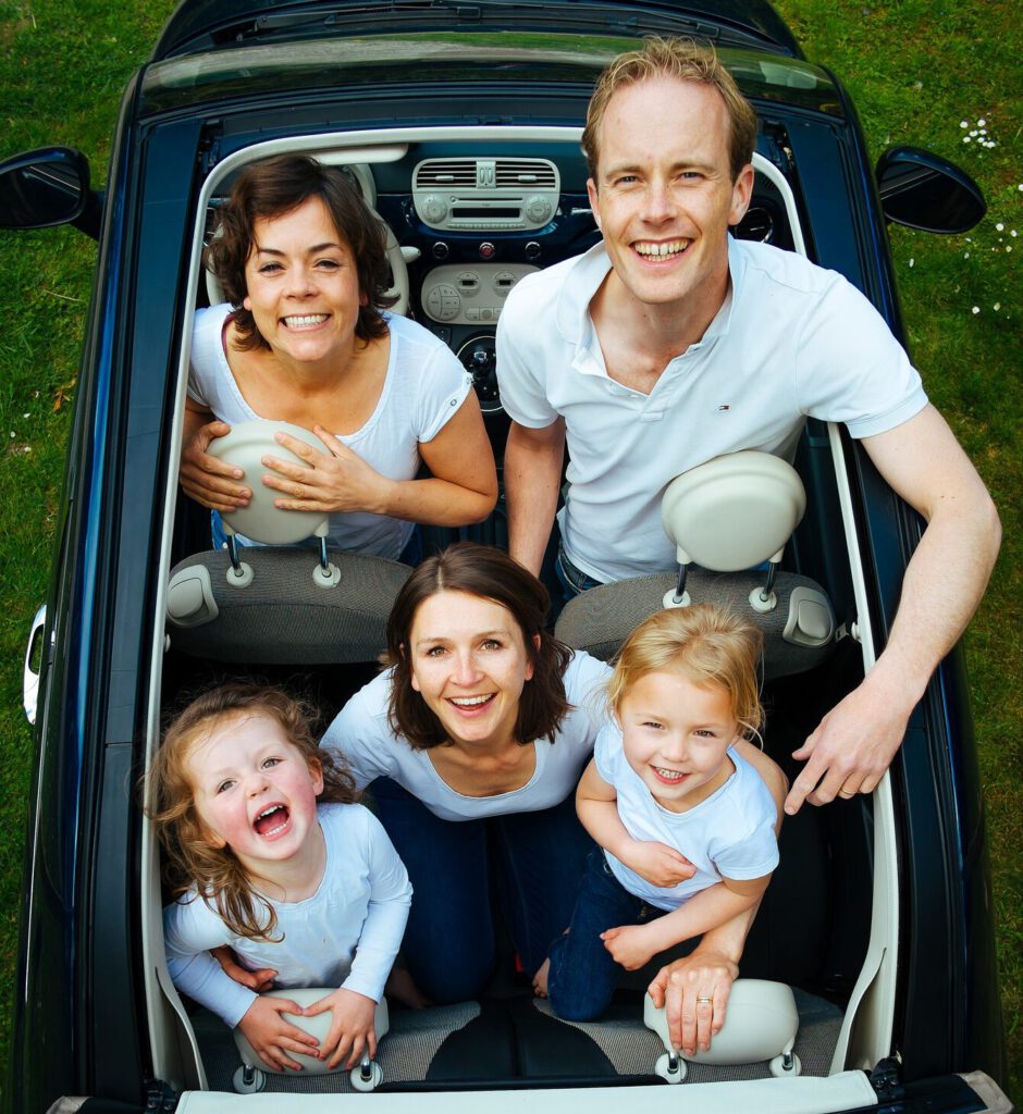 L wie lange Fahrten. Familie sitzt im Auto und lächelt.